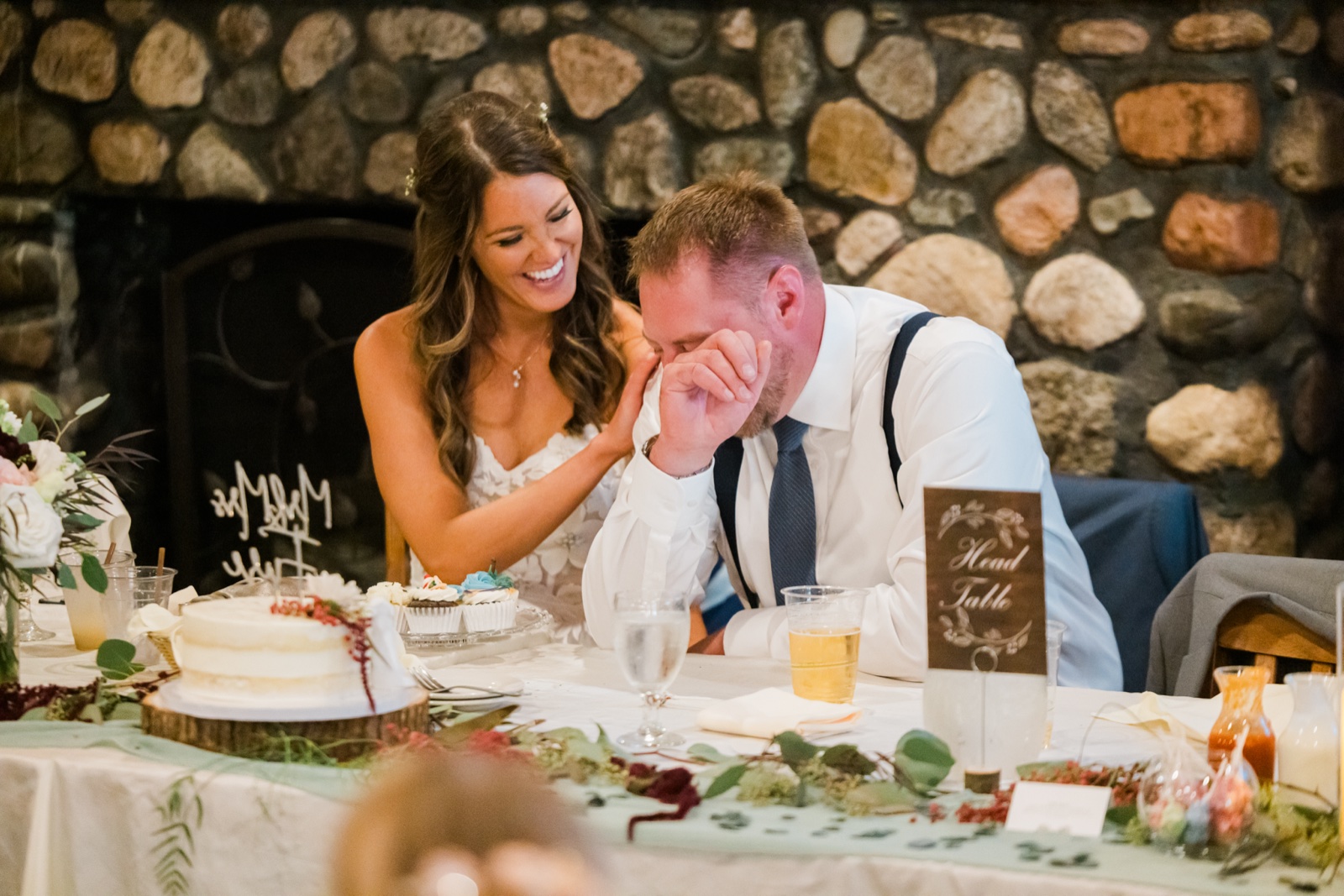 Bride laughs while the groom wipes tears during toasts at the head table by the stone fireplace at Manhattan Beach Lodge — Tim Larsen Photography, Brainerd Lakes MN