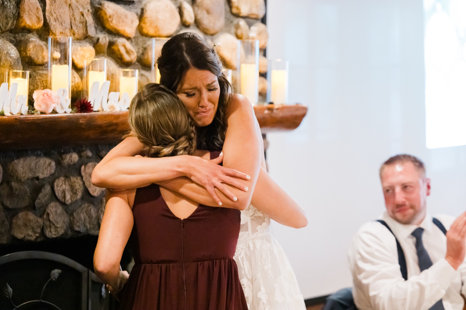 Bride hugs a bridesmaid after toasts by the candle-lined stone fireplace at Manhattan Beach Lodge — Tim Larsen Photography, Brainerd Lakes MN