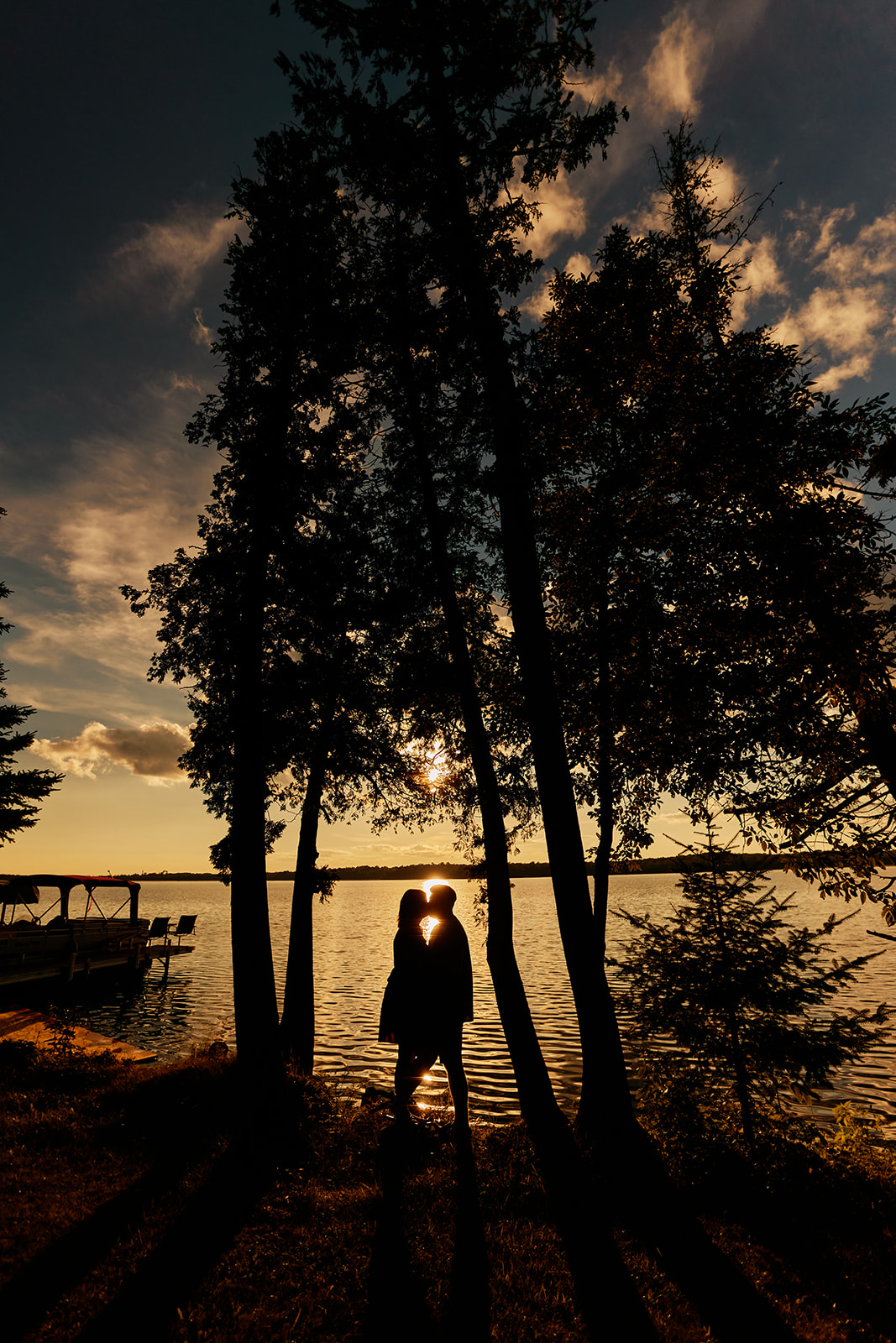 A silhouetted couple stands embracing between two tall pine trees with the sun bursting between them over a Minnesota lake — Tim Larsen Photography, Brainerd Lakes MN