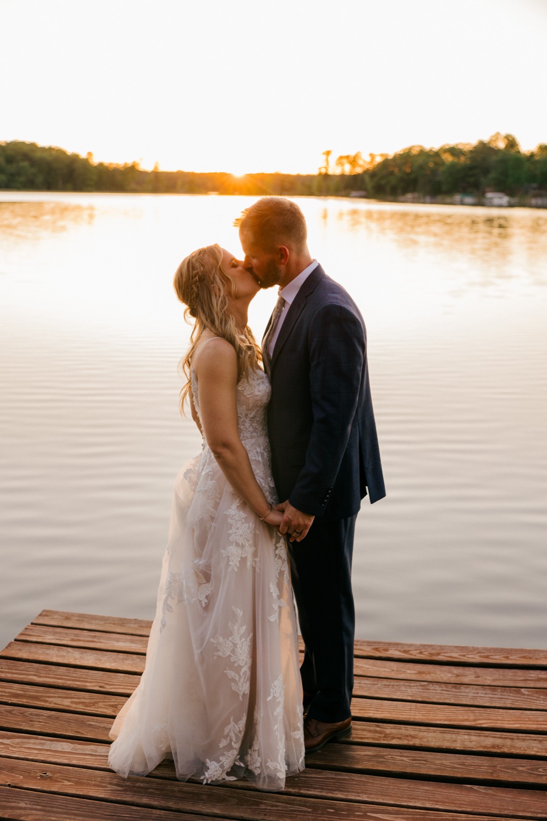 A couple kisses at the end of a wooden lakeside dock as the sun flares behind them at golden hour over a private Minnesota lake — Tim Larsen Photography, Brainerd Lakes MN