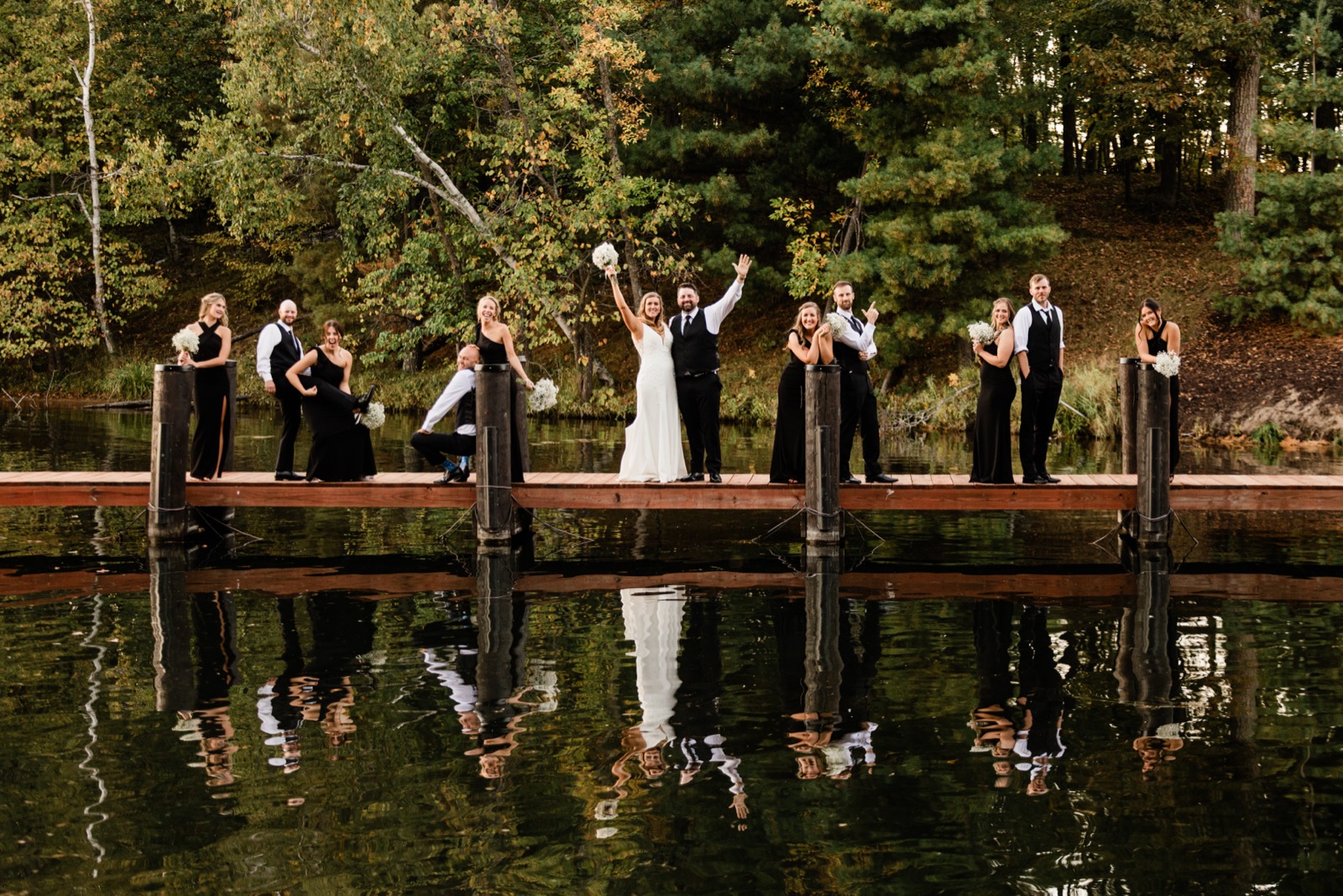A bridal party of nine spread across a long lakeside dock with a perfect mirrored reflection in the still water of Whitefish Bay — Tim Larsen Photography, Brainerd Lakes MN