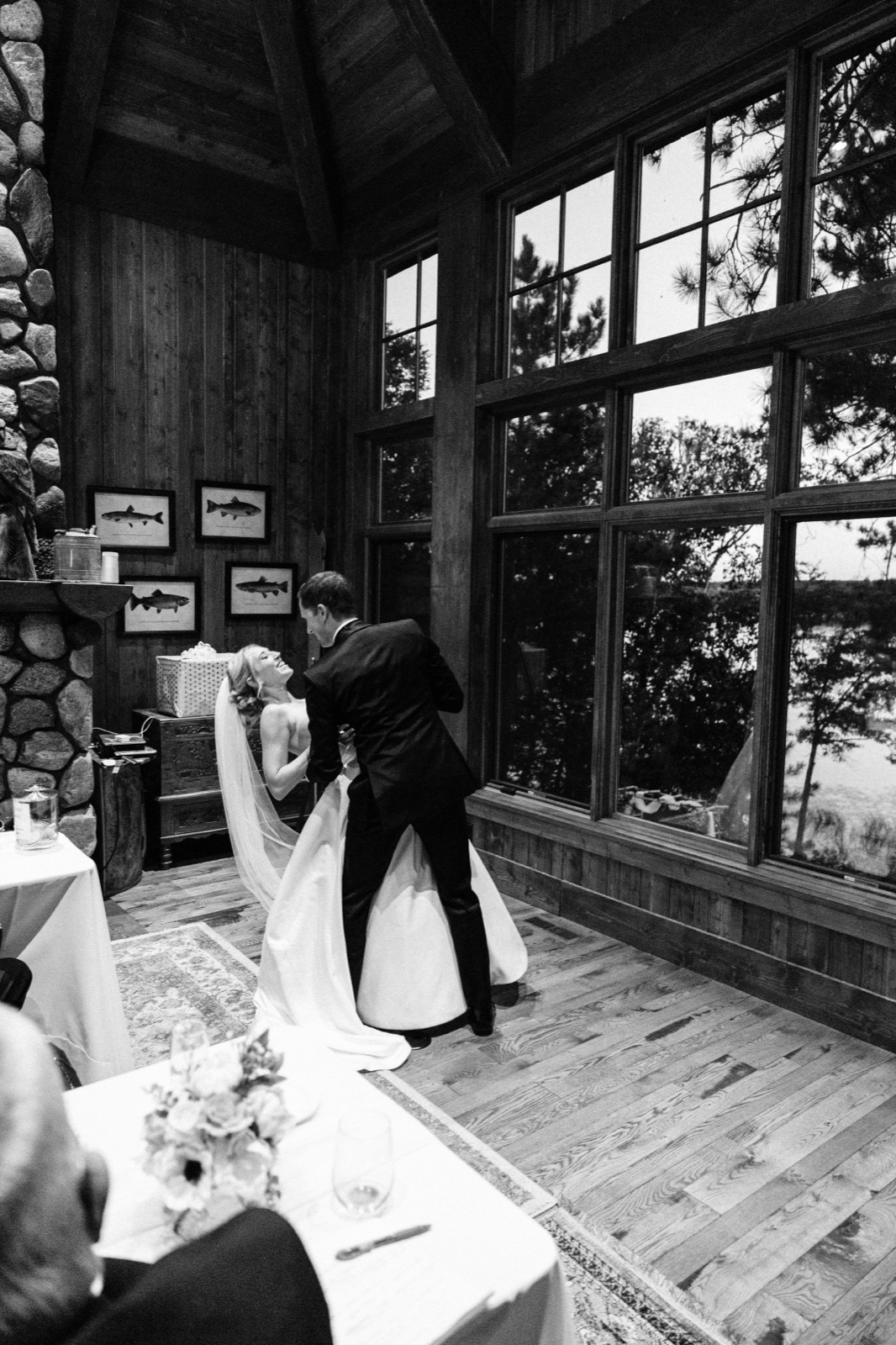 A bride throws her head back laughing as the groom dips her in front of a stone fireplace inside a private lake cabin great room — Tim Larsen Photography, Brainerd Lakes MN