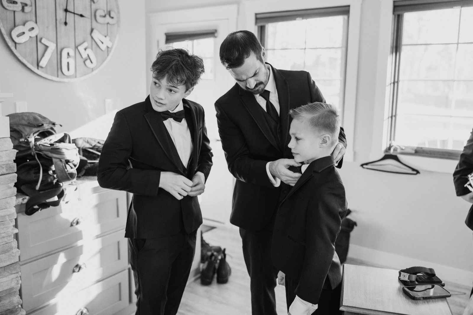 In black and white, the groom adjusts the collar of a young boy while an older boy buttons his own tuxedo jacket in a window-lit room at Grand View Lodge — Tim Larsen Photography, Brainerd Lakes MN