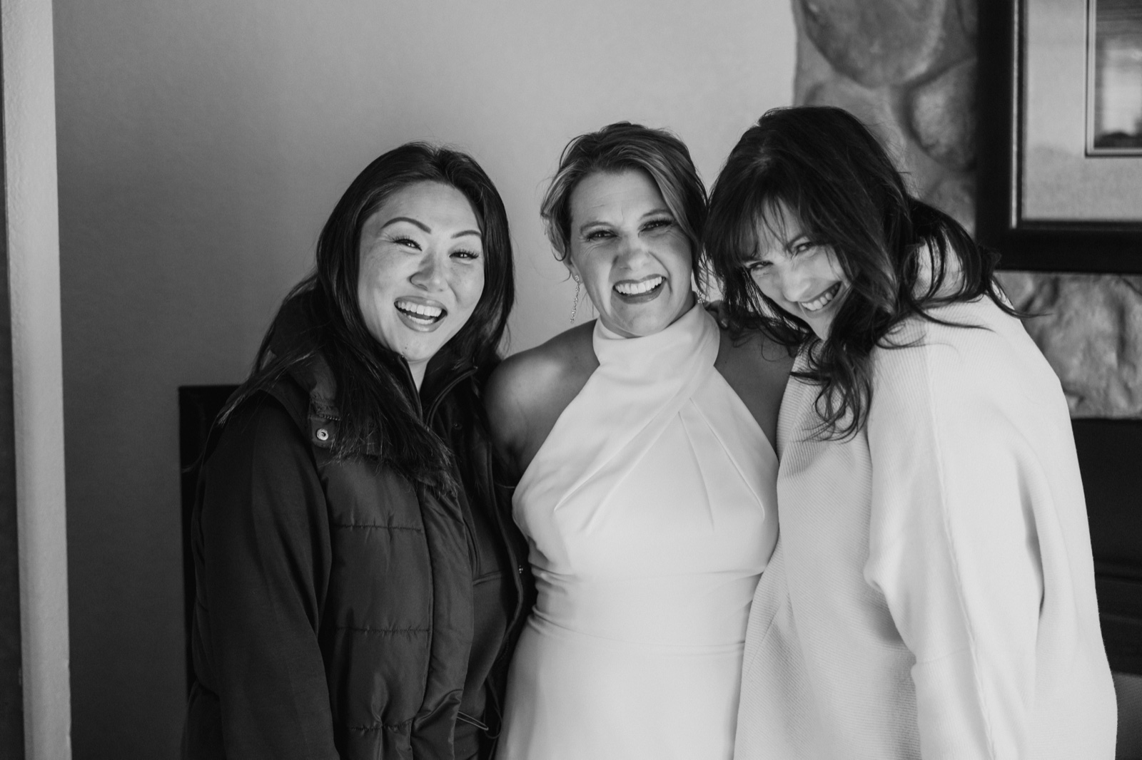The bride laughs in the center while two friends lean in on either side of her, all three caught mid-laughter in a window-lit room.