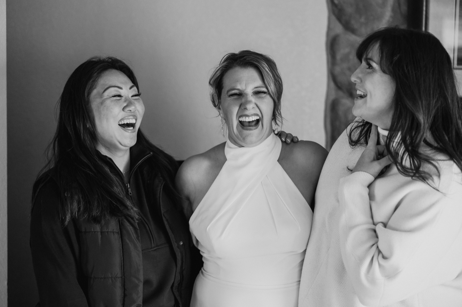 Black-and-white image of the bride in a halter gown laughing openly between two friends who are also mid-laugh in a getting-ready room.