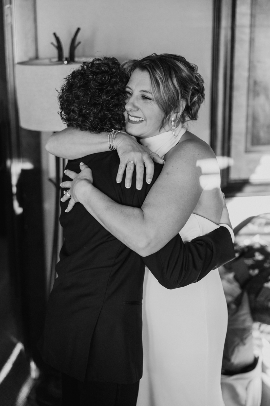 The bride smiles with visible emotion as she embraces a loved one in a window-lit room, rendered in black and white — Tim Larsen Photography, Brainerd Lakes MN