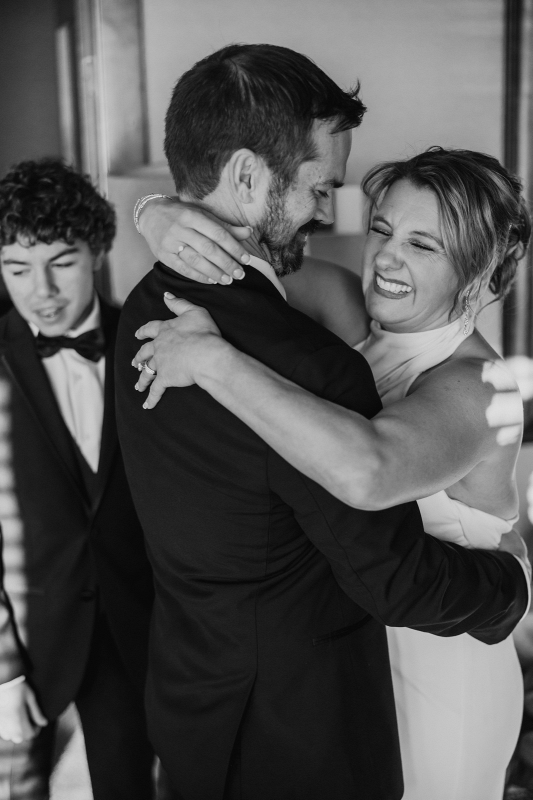 The bride laughs with her eyes closed while embracing the groom, with a young guest in a tuxedo visible in the foreground.