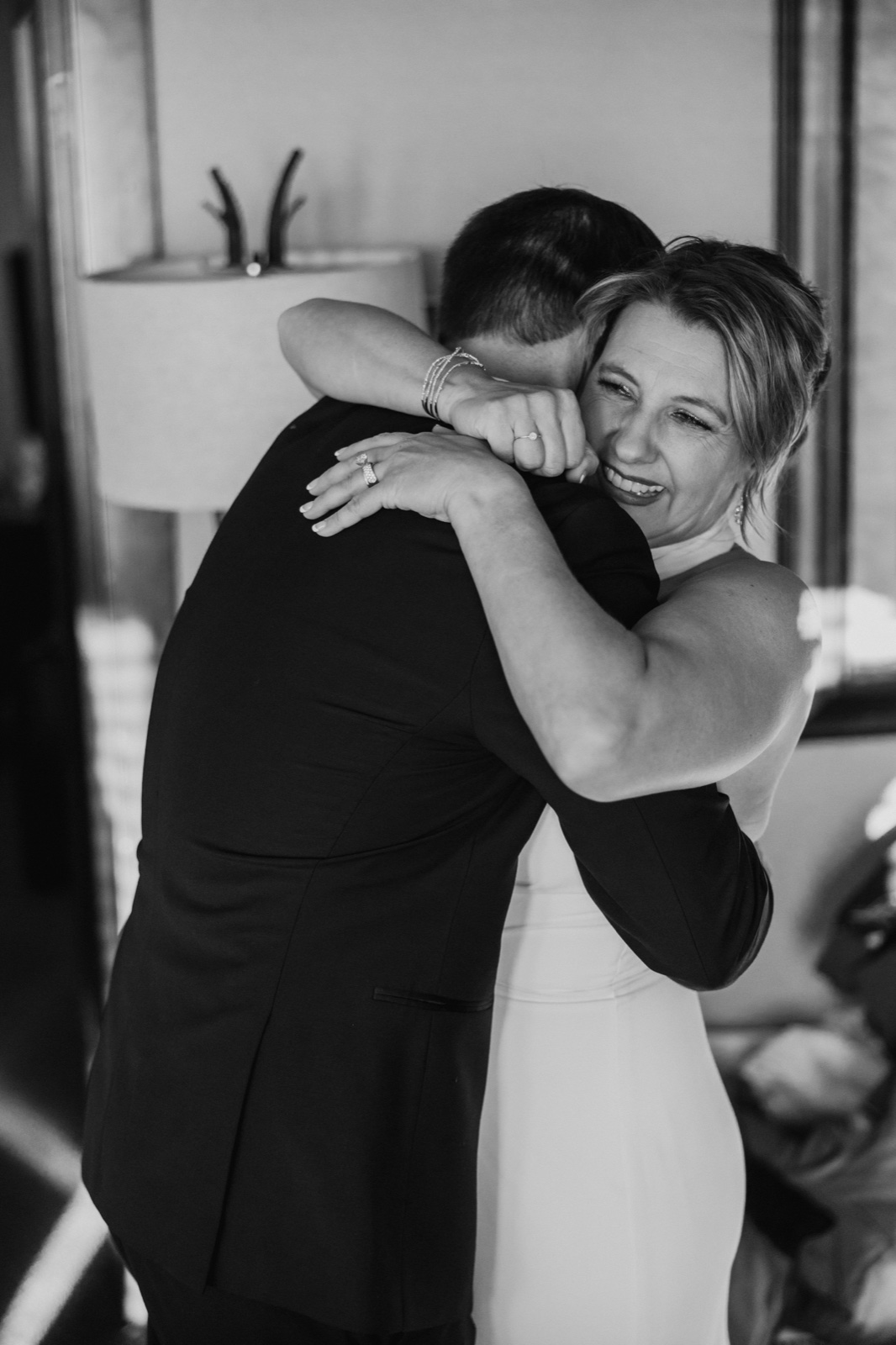 The bride smiles wide with eyes closed as she wraps her arms around the groom in a tight embrace during their first look in a window-lit room.