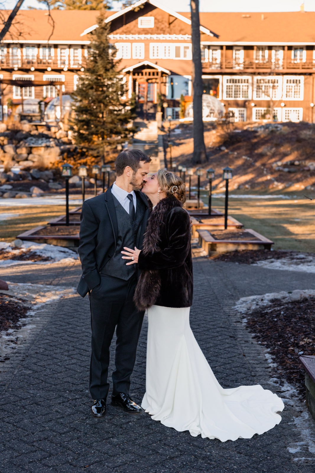 The bride in a white gown and fur stole kisses the groom in a black suit on a paver path, with the illuminated wood facade of Grand View Lodge glowing behind them in winter afternoon light.