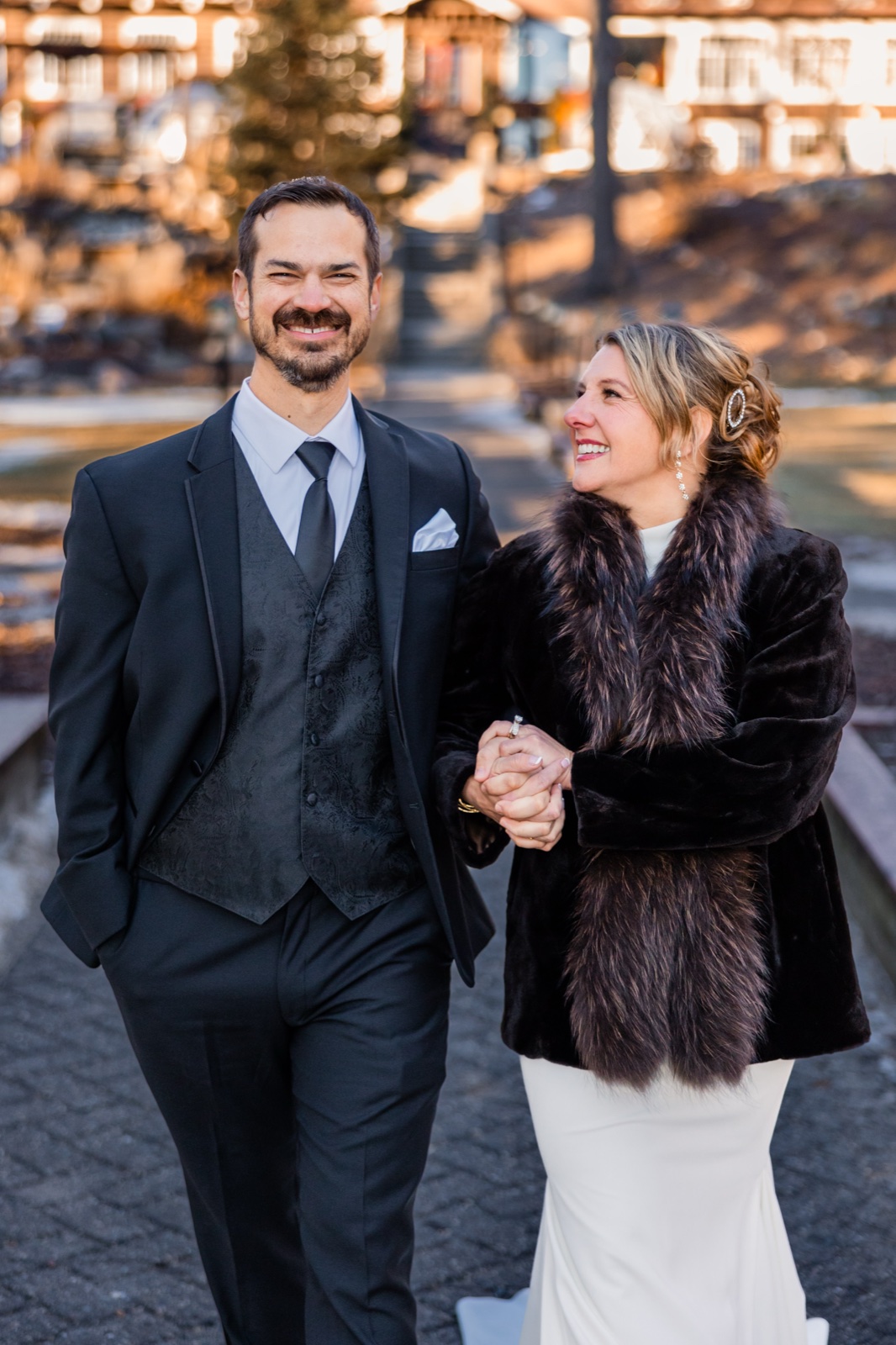 The groom laughs toward the camera while the bride, wrapped in a dark fur stole, looks at him smiling as they walk hand in hand outside Grand View Lodge in warm afternoon light.