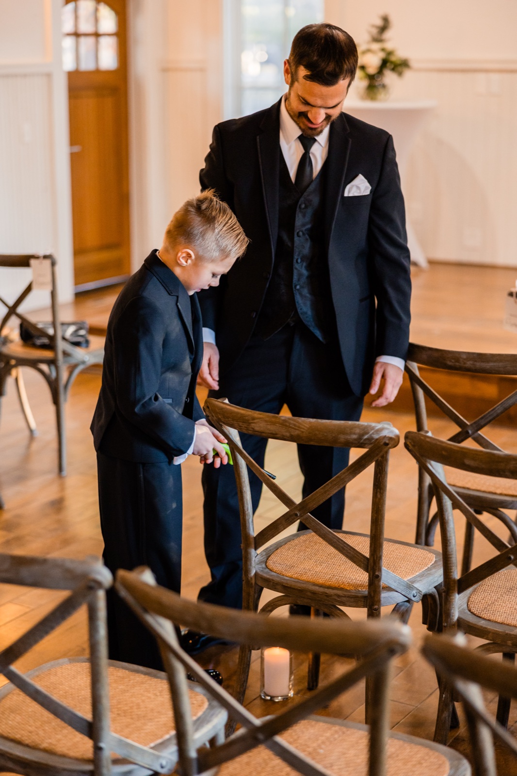 The groom stands with a young boy in a matching suit who looks down at something in his hands, framed by wooden cross-back chairs in the ceremony room at Grand View Lodge — Tim Larsen Photography, Brainerd Lakes MN