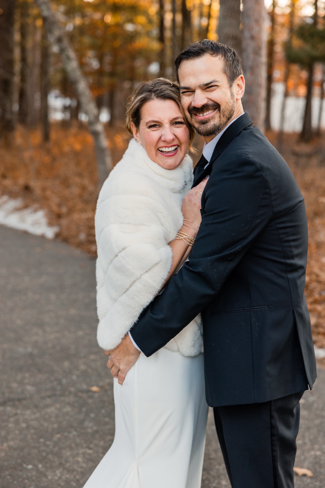 The bride in a white fur stole and the groom in a dark suit laugh together, cheek-to-cheek, on a wooded path with backlit winter trees behind them at Grand View Lodge.