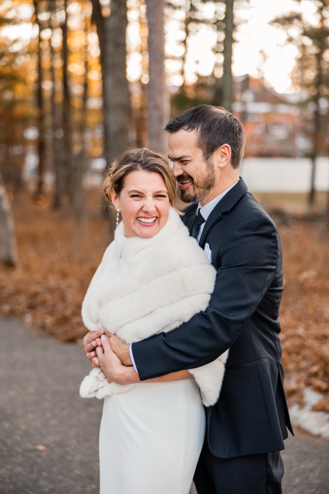 The bride laughs broadly toward the camera wrapped in a white fur stole as the groom embraces her from behind and leans his face into her hair on a wooded path at Grand View Lodge.