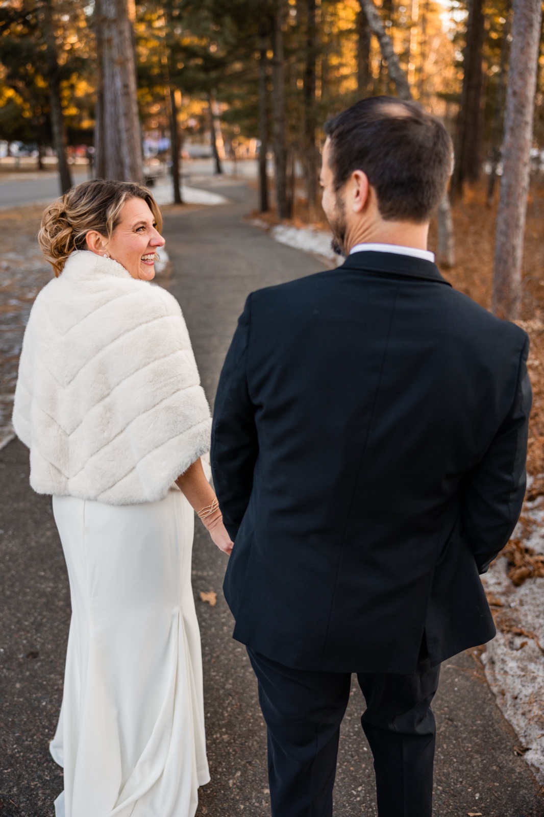 The bride, wearing a white fur stole over her gown, glances back laughing at the groom as they walk down a tree-lined path in warm evening light at Grand View Lodge.