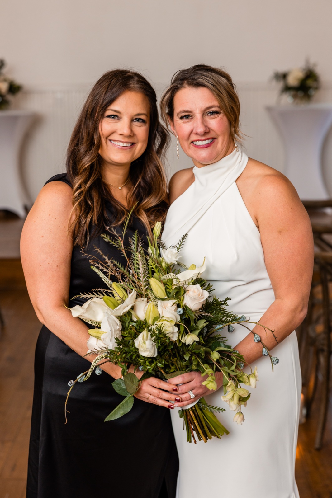 The bride in a white halter gown stands beside a bridesmaid in a black dress, both smiling at the camera while the bride holds a large greenery-and-white bouquet.