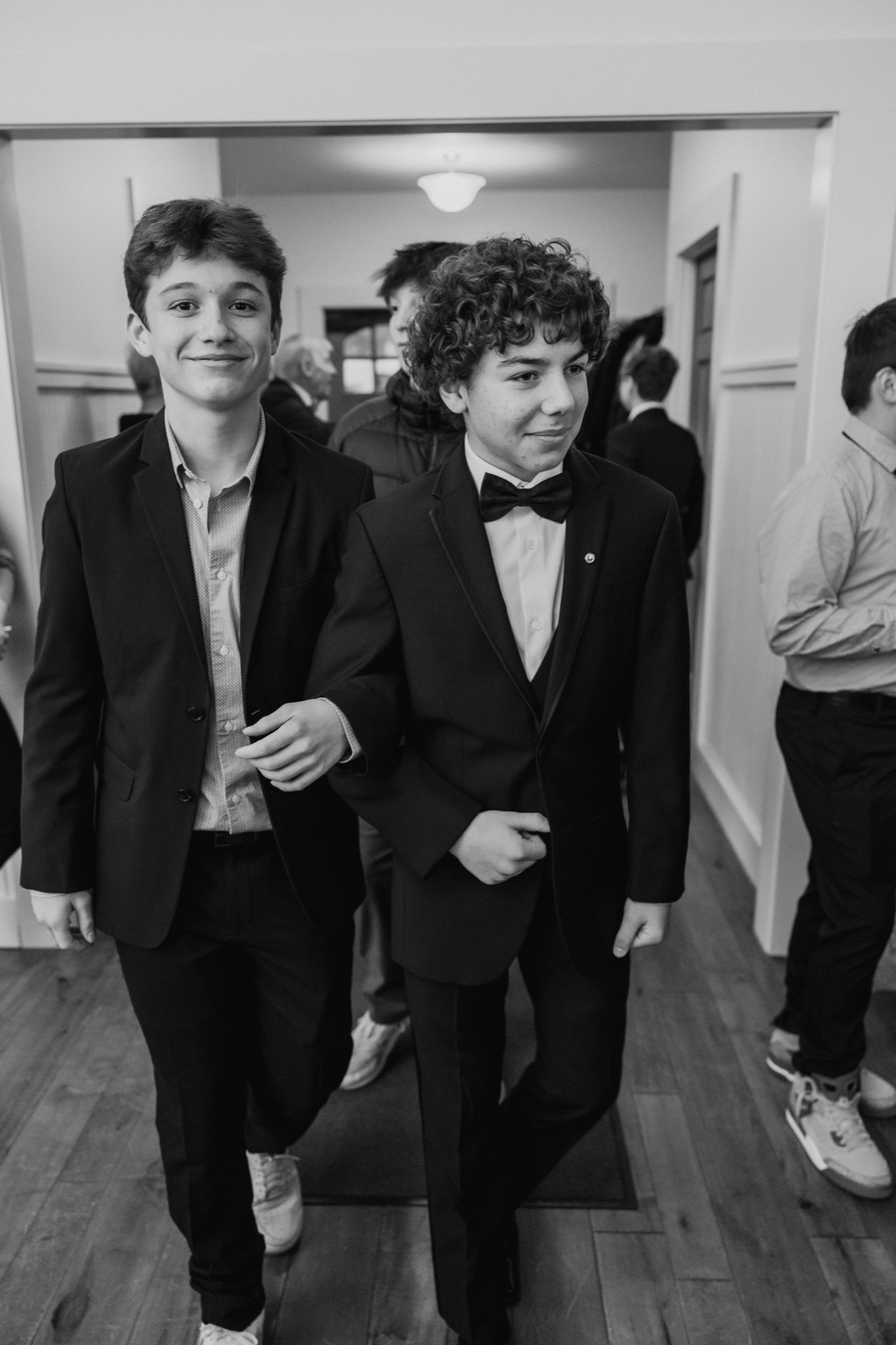 Two young guests in tuxedos walk together through a doorway at Grand View Lodge while other guests mingle in the hallway behind them — Tim Larsen Photography, Brainerd Lakes MN