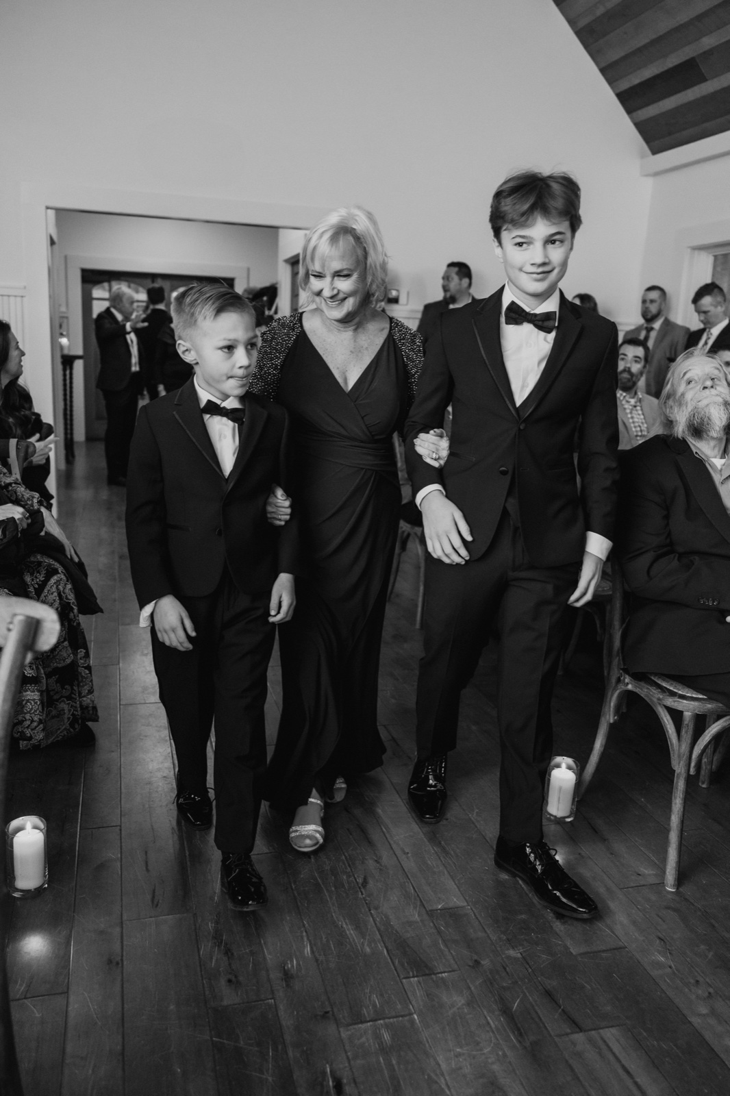 A mother in a dark gown walks down a candle-lined aisle flanked by her two sons in tuxedos, smiling down as seated guests watch at Grand View Lodge.