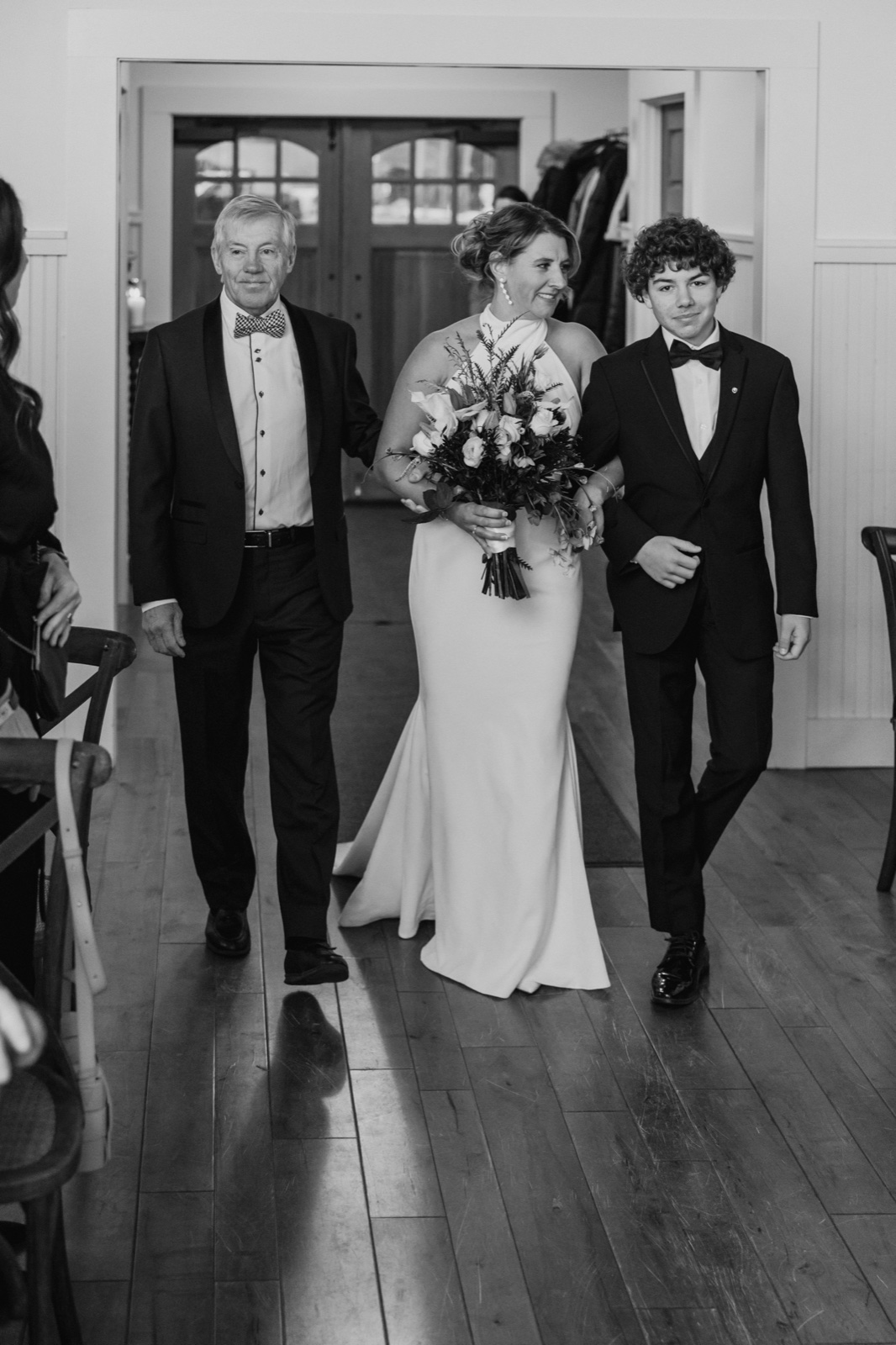 The bride walks down the aisle flanked by her father and son, framed in the open doorway of the Grand View Lodge ceremony space.