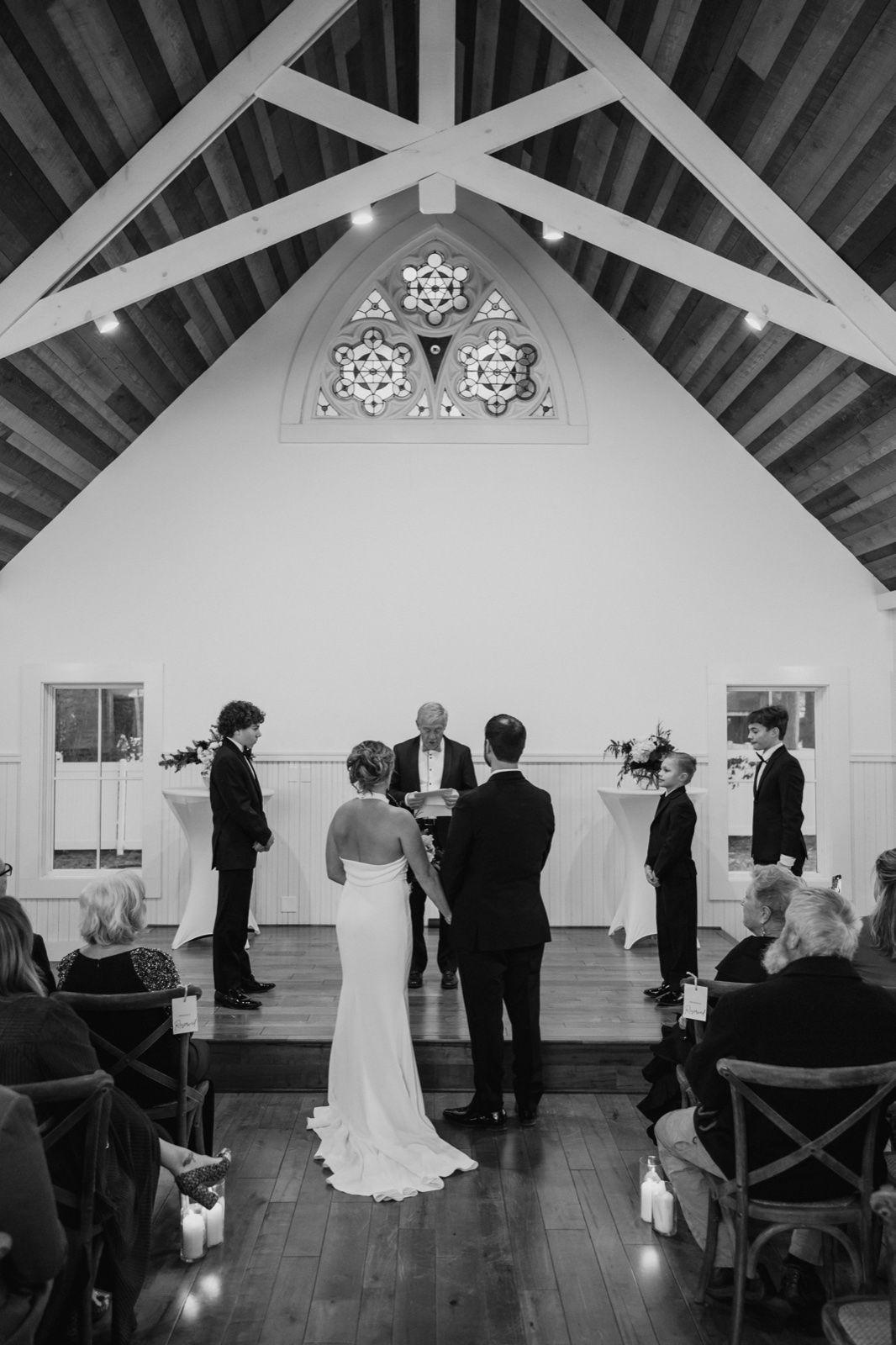 A wide black-and-white view of the bride and groom holding hands at the altar beneath a vaulted wood-beam ceiling and a rose stained-glass window at Grand View Lodge.