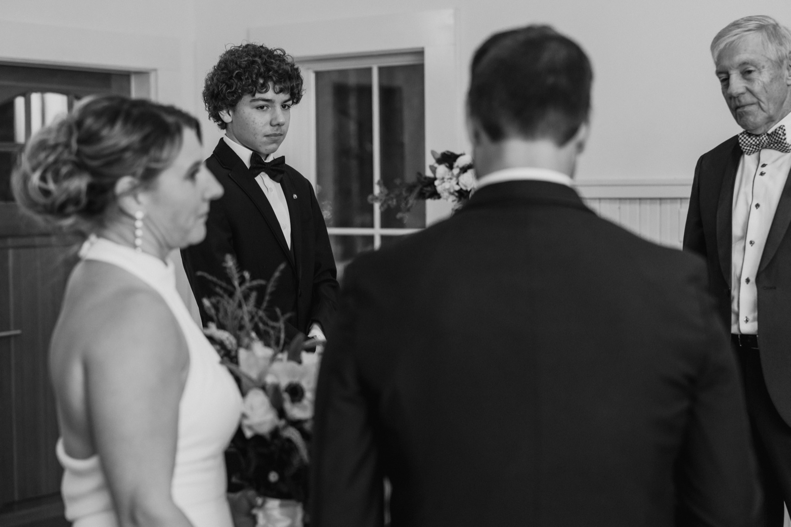 A young guest in a tuxedo watches intently between the soft-focused figures of the bride and groom during an indoor ceremony.