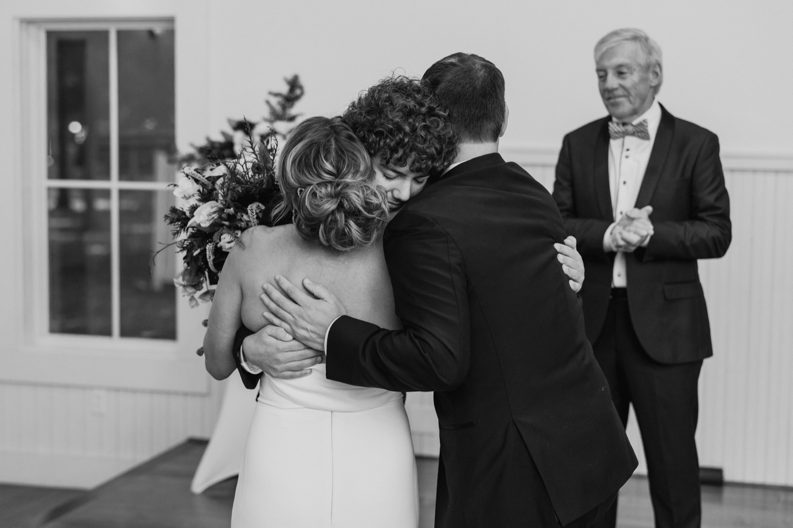 The bride embraces a family member in a three-way hug while her father watches from the background with clasped hands, rendered in black and white — Tim Larsen Photography, Brainerd Lakes MN