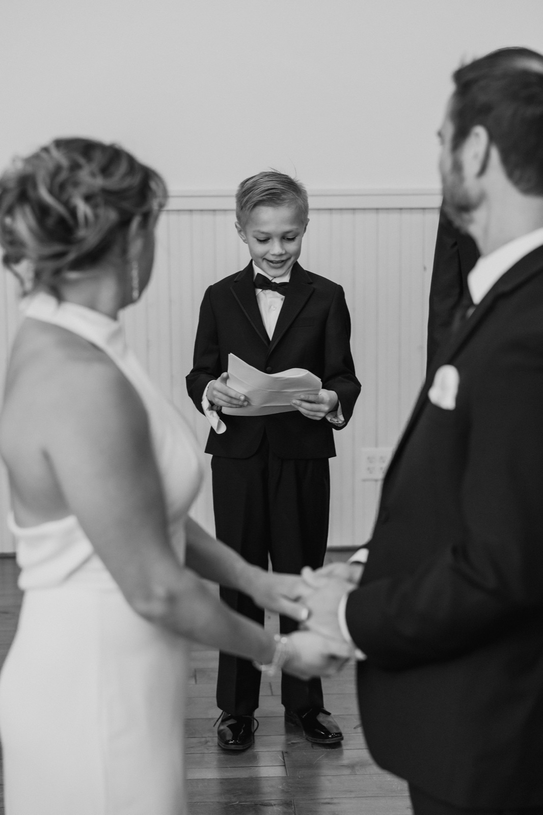 A young boy in a black tuxedo reads from a folded paper while standing between the out-of-focus bride and groom during the ceremony — Tim Larsen Photography, Brainerd Lakes MN