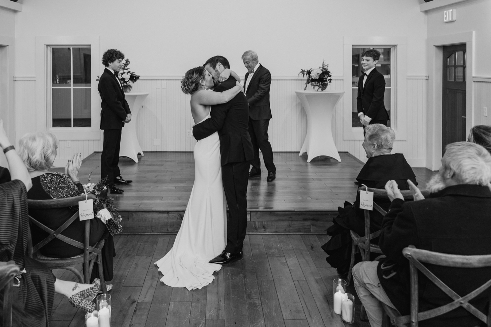 The bride and groom share their first kiss at the center of an intimate ceremony room while the officiant, a young attendant, and seated guests look on — Tim Larsen Photography, Brainerd Lakes MN