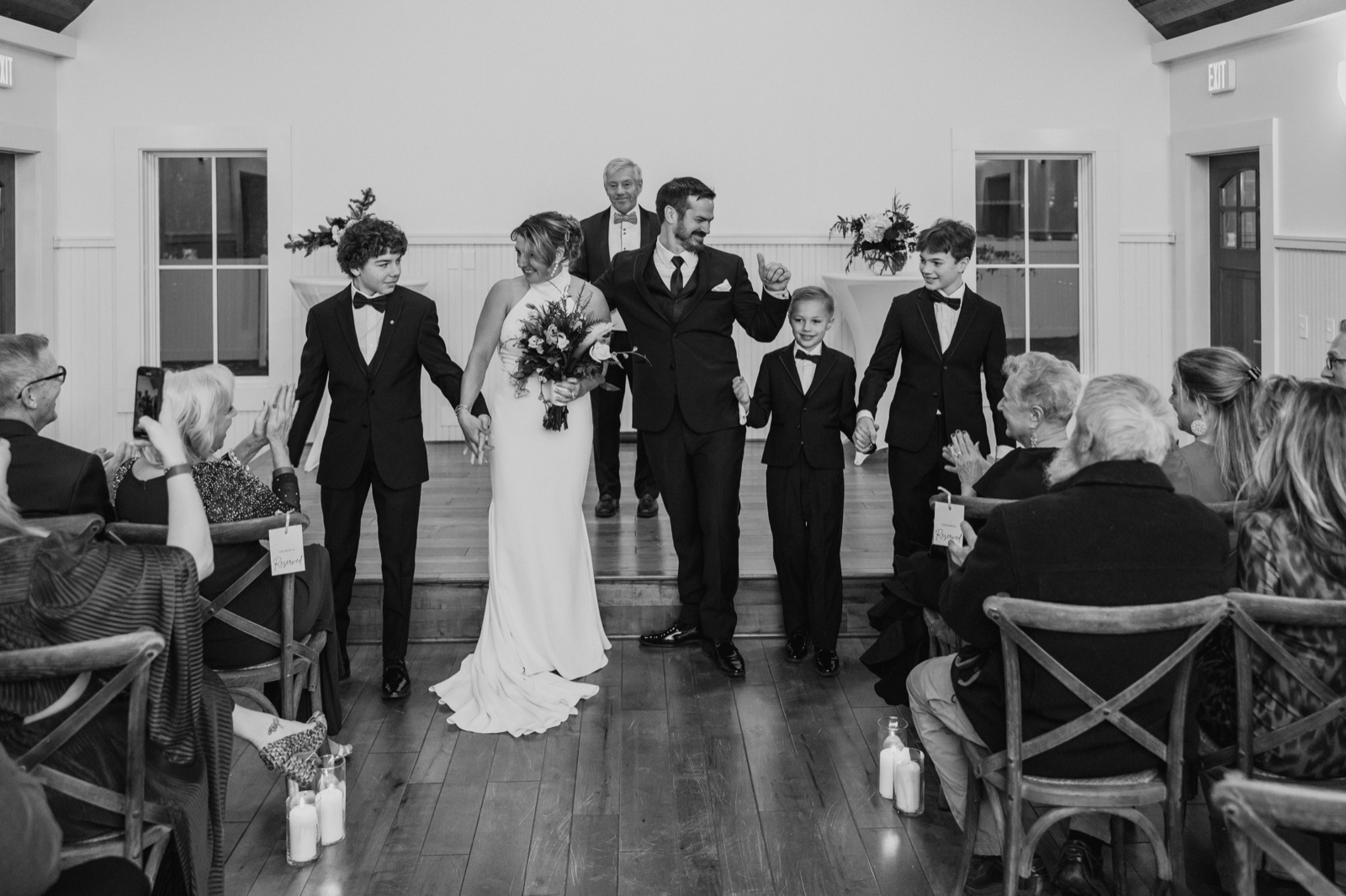 The bride and groom walk back up the aisle hand-in-hand with three boys beside them as seated guests clap from both sides of the ceremony room.