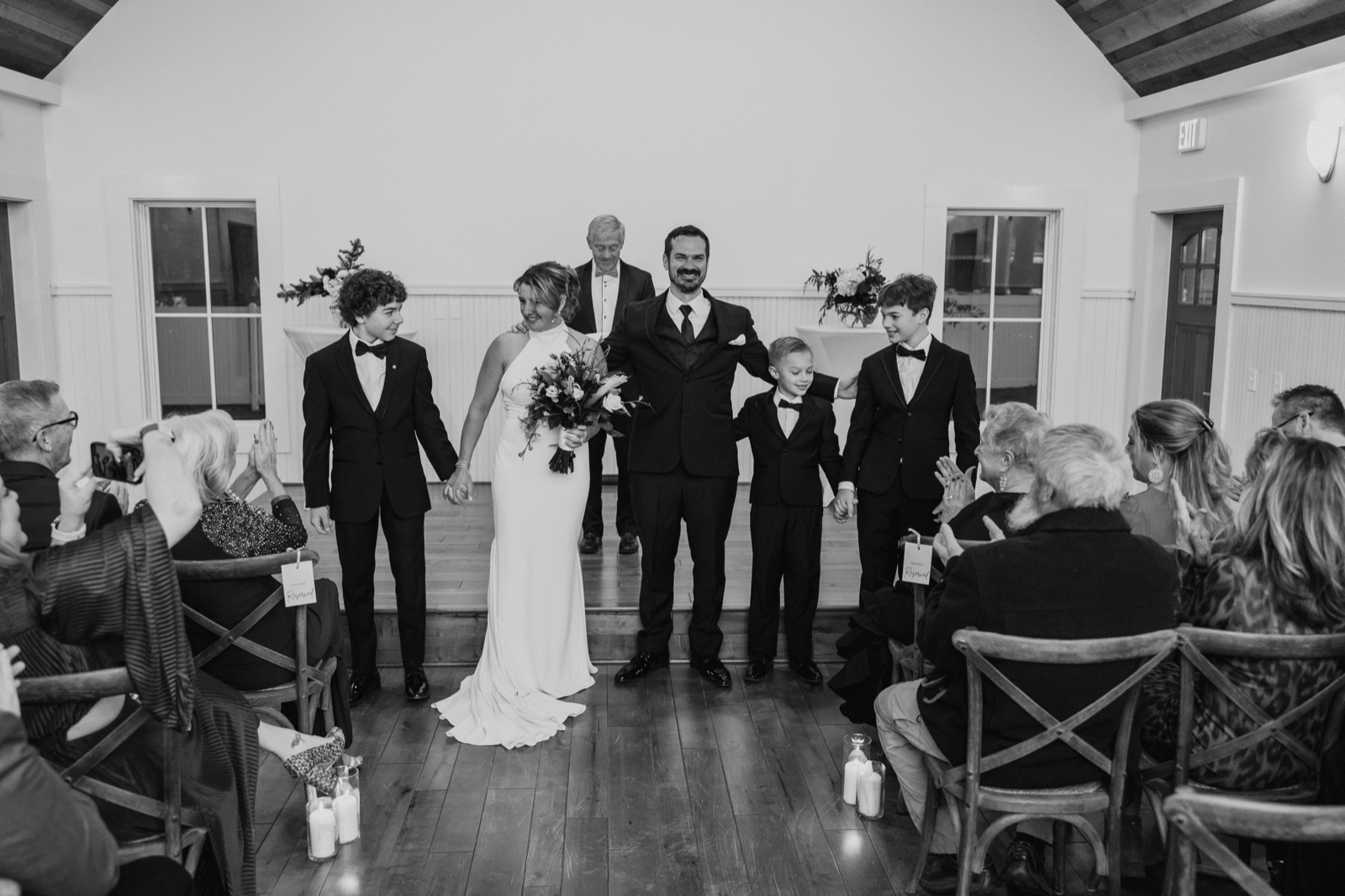 The bride and groom stand at the front of the ceremony room flanked by their sons as guests applaud from chairs lining the aisle at Grand View Lodge.