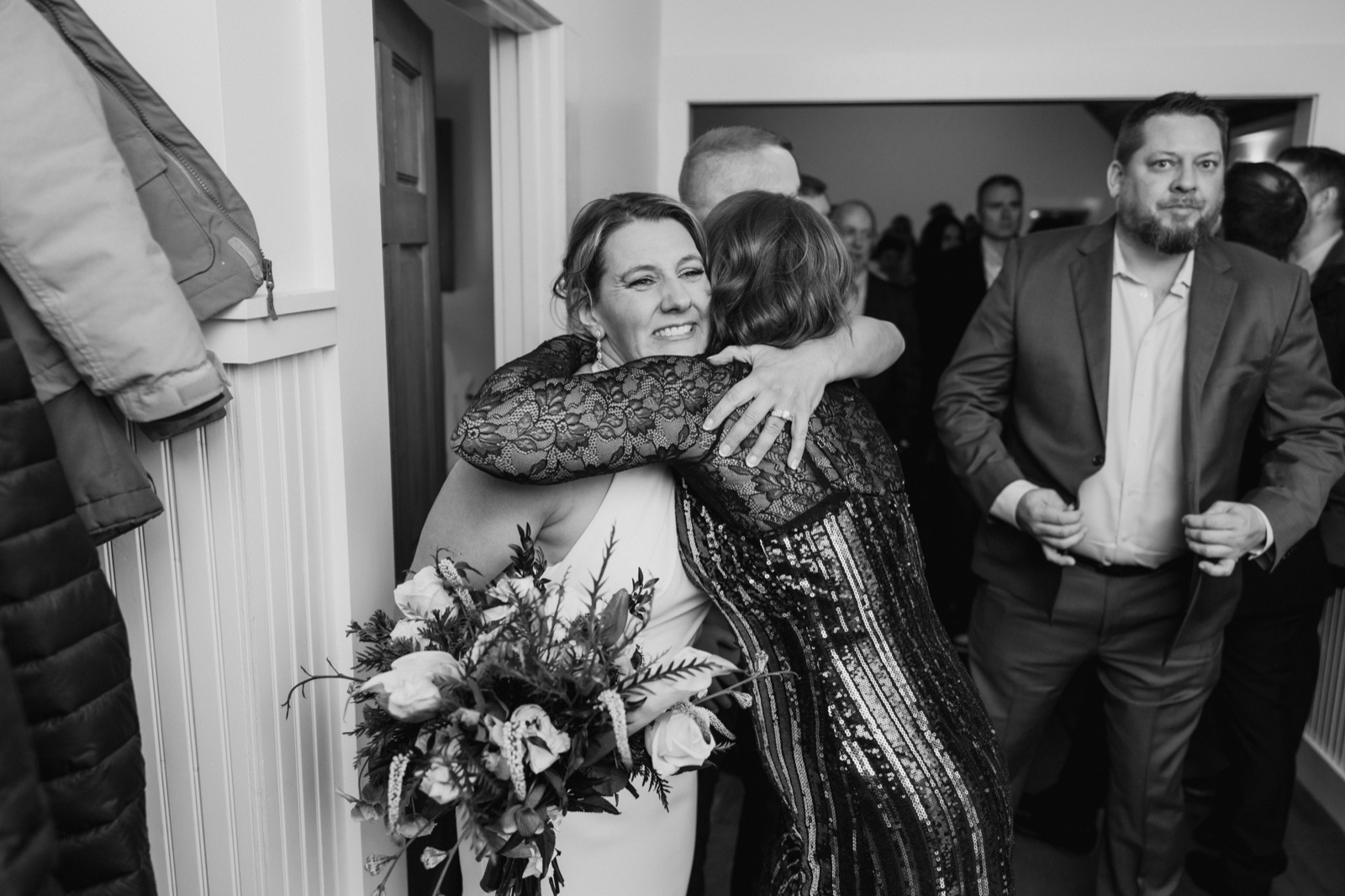 In black and white, the bride smiles broadly as she embraces a guest in a sequined gown in a hallway while another guest watches from the right.