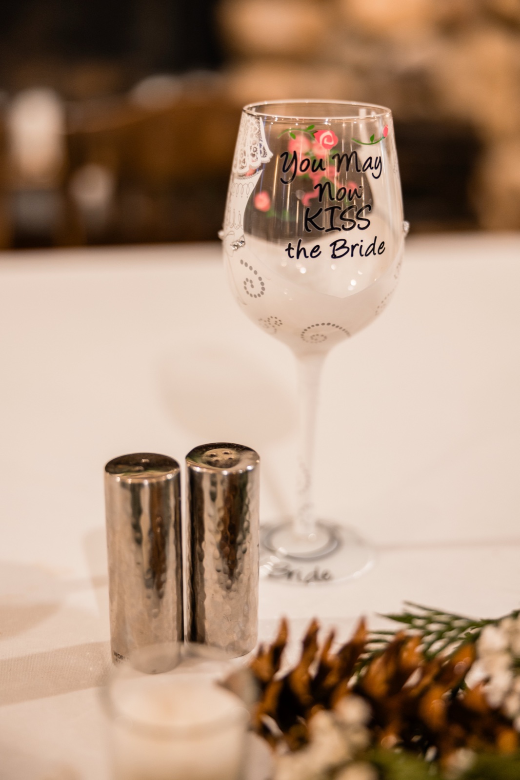 A wine glass printed with "You May Now KISS the Bride" sits on a reception table beside hammered silver salt and pepper shakers and a pinecone-and-baby's-breath centerpiece.