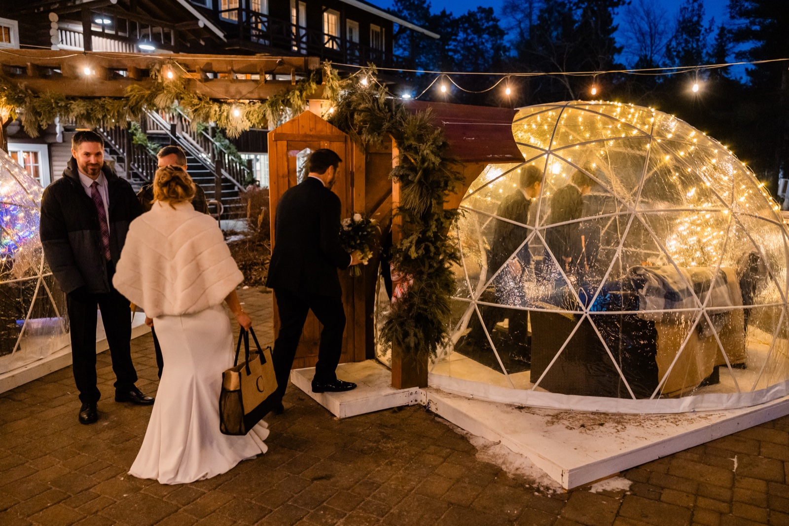 The bride in a white fur wrap walks with the groom toward a string-lit igloo dome outside Grand View Lodge at dusk, guests gathered nearby.