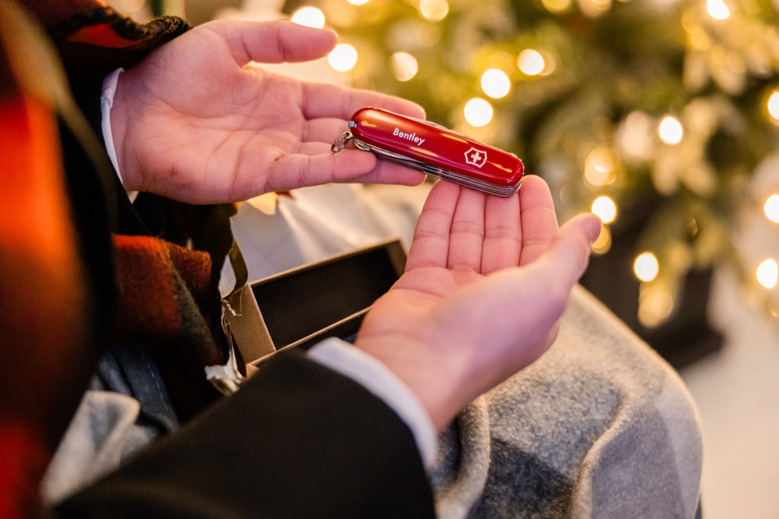 A personalized red Swiss Army knife engraved with the name Bentley rests in a groomsman's open hands in front of a lit Christmas tree.