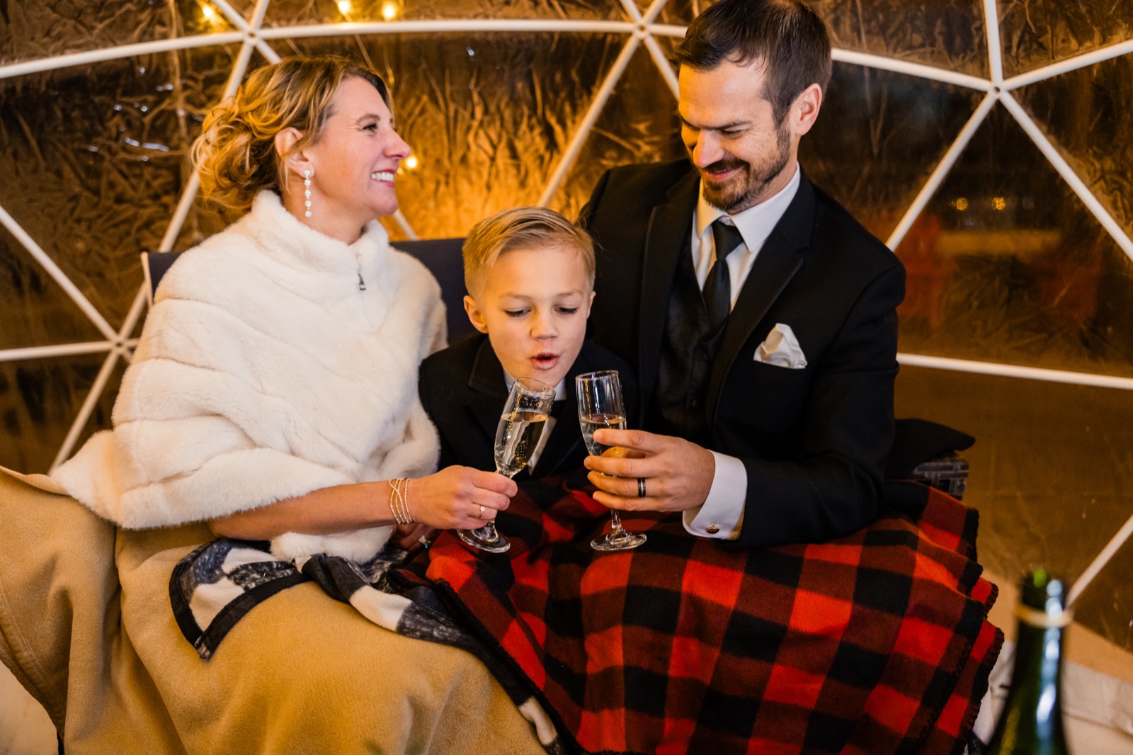 The bride in a white fur wrap laughs as a young boy in a suit puckers his lips toward a champagne flute while the groom watches, all three seated under a lit geodesic dome with a plaid blanket across their laps at Grand View Lodge.