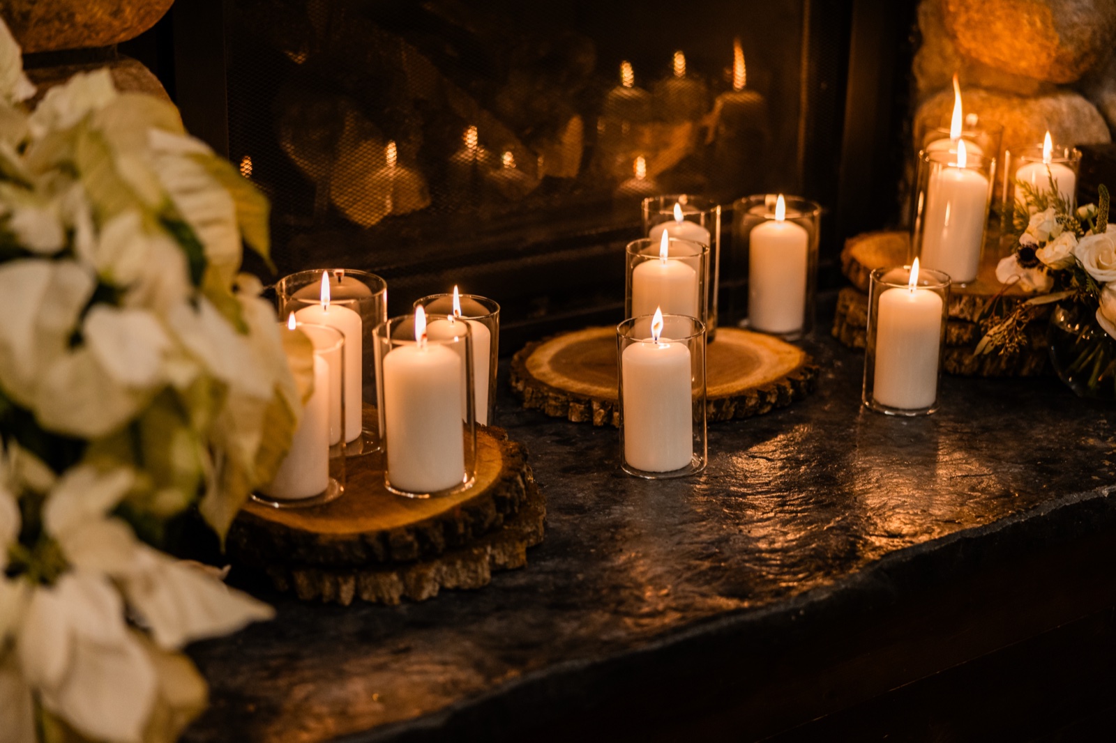 Pillar candles in glass holders rest on wood slab rounds along a stone hearth in front of a lit fireplace, with white poinsettias framing the left side.
