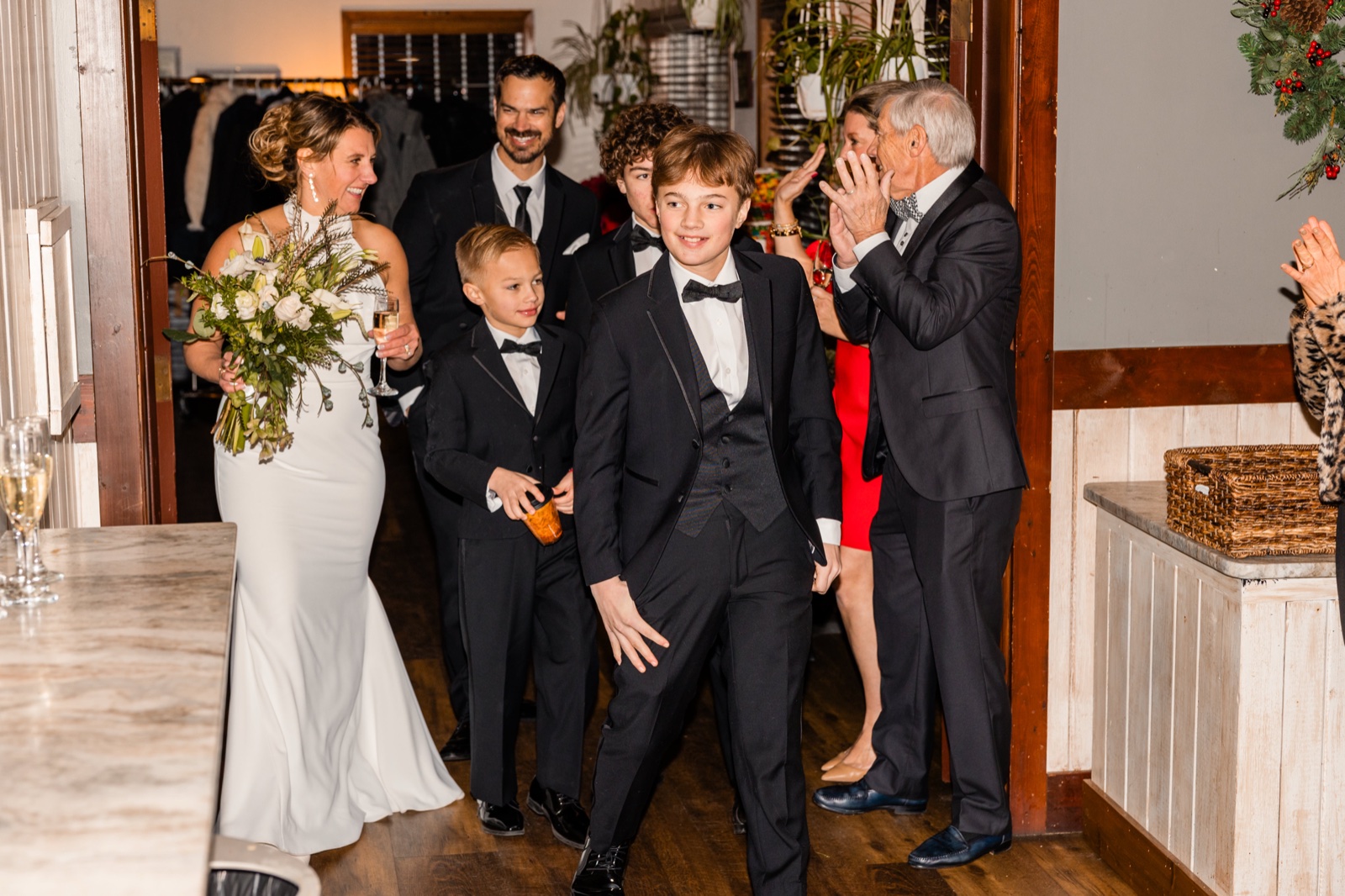 A young boy in a tuxedo leads the bride and family into the reception as an older guest claps in reaction beside them.
