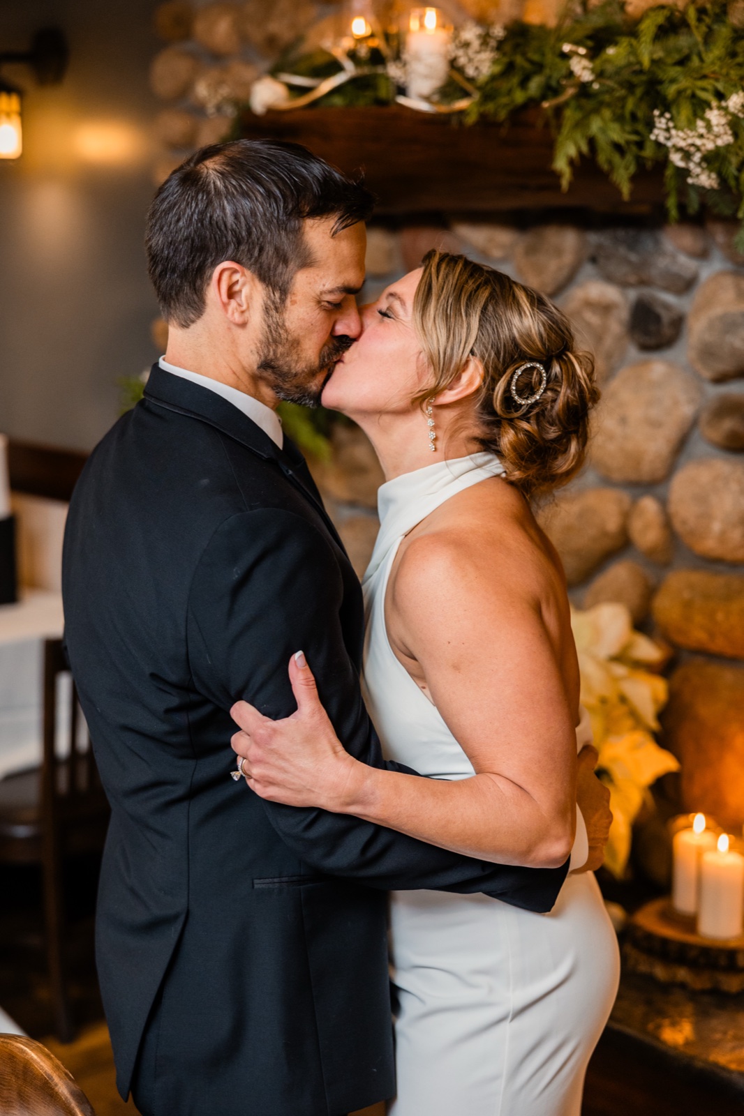 The bride and groom share a kiss in front of a stone fireplace decorated with evergreens and lit candles at Grand View Lodge.