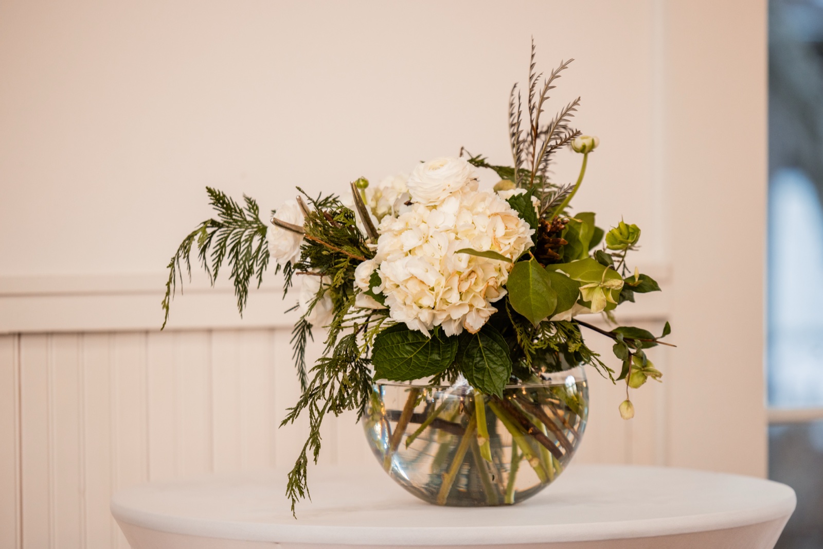 A glass bowl centerpiece with white hydrangeas, ranunculus, cedar, and trailing greenery sits on a linen-draped table against a white beadboard wall at Grand View Lodge.