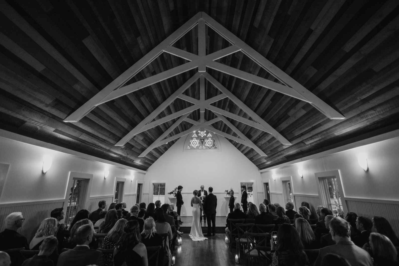 A black-and-white wide-angle view of a wedding ceremony inside a chapel with a dramatic vaulted wooden truss ceiling, the couple standing at the altar with their officiant as seated guests fill the pews.