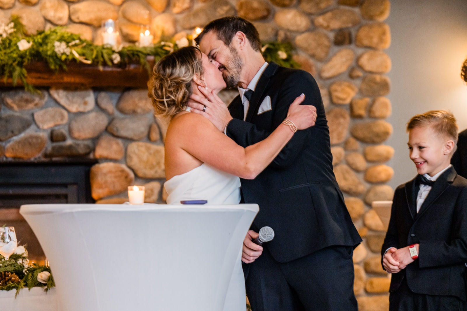 The bride and groom share a kiss in front of a stone fireplace draped with greenery while a young boy in a tuxedo grins at the moment from the right — Tim Larsen Photography, Brainerd Lakes MN