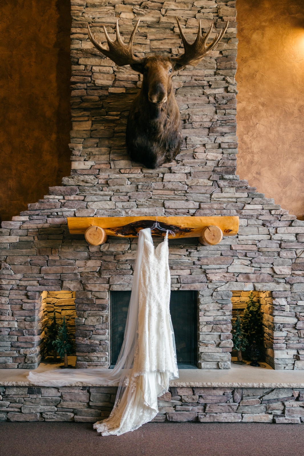 The bride's lace wedding dress with a long veil hangs from the log mantel of a stacked-stone fireplace beneath a mounted moose head at Whitefish Lodge — Tim Larsen Photography, Brainerd Lakes MN