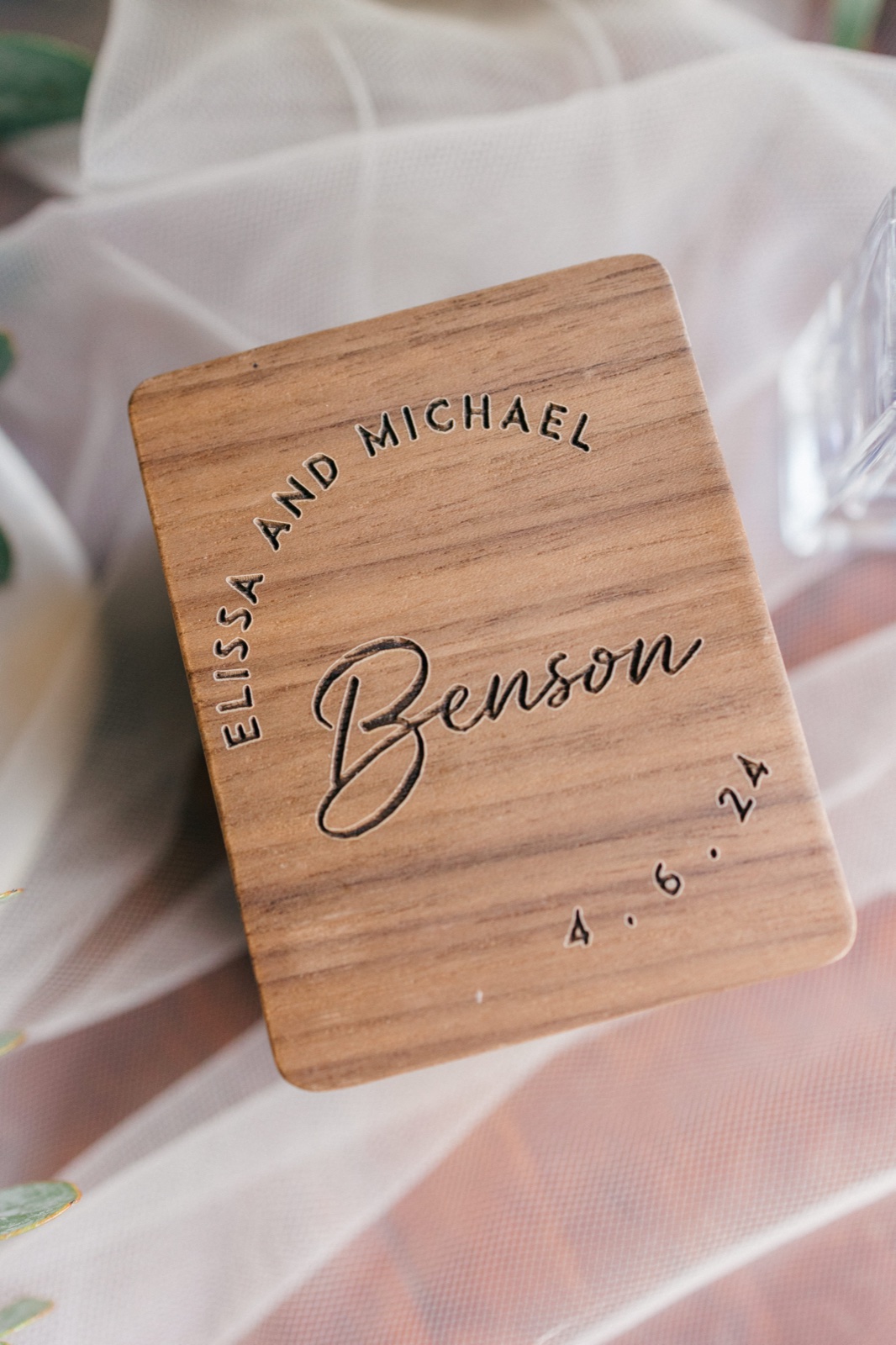 A wooden ring box engraved with the couple's names rests on a soft white veil with greenery at the edges — Tim Larsen Photography, Brainerd Lakes MN