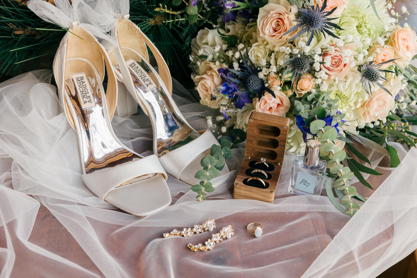 A flat-lay of the bride's Badgley Mischka heels, wedding rings in a wooden box, pearl earrings, an opal ring, a perfume bottle, and a peach and blue thistle bouquet on a tulle veil — Tim Larsen Photography, Brainerd Lakes MN