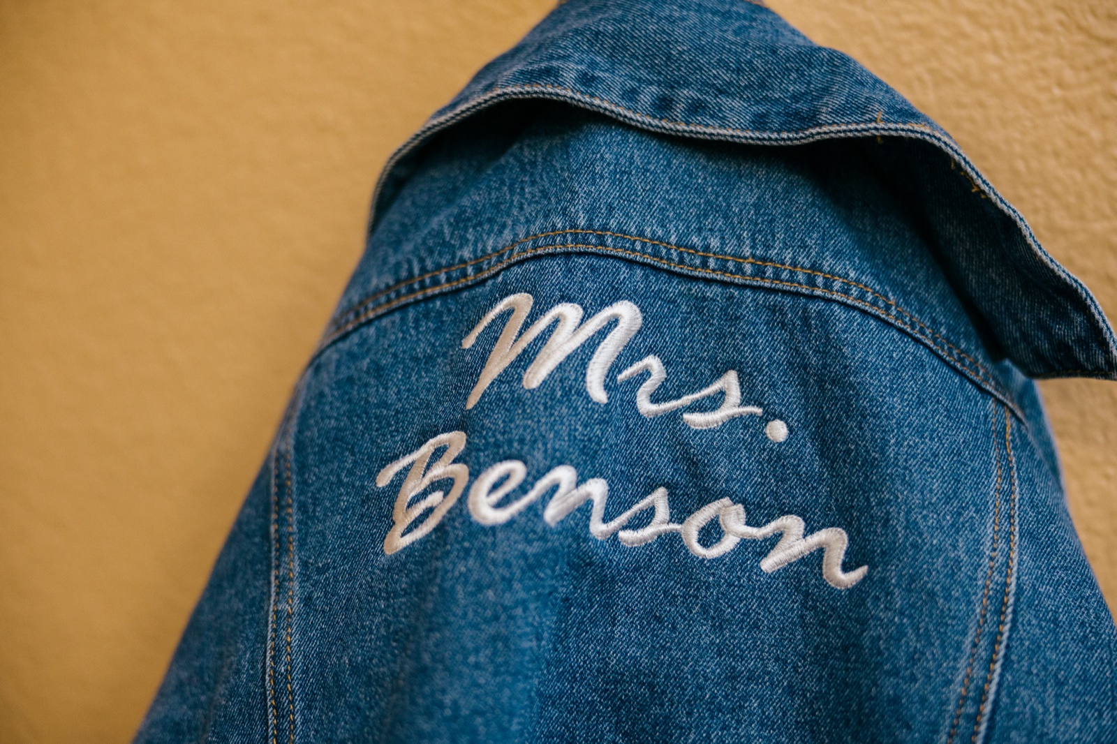 A denim jacket with 'Mrs. Benson' embroidered in white cursive across the back, hung against a warm yellow wall — Tim Larsen Photography, Brainerd Lakes MN
