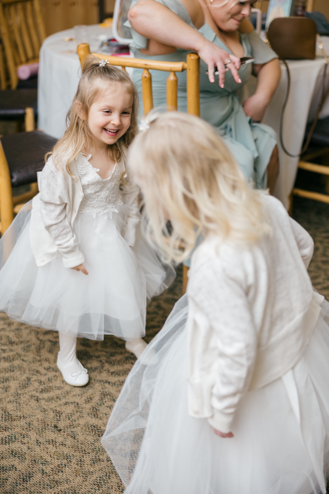 A young flower girl in a white tulle dress laughs brightly while a bridesmaid in sage green sits in the background of the getting-ready room — Tim Larsen Photography, Brainerd Lakes MN