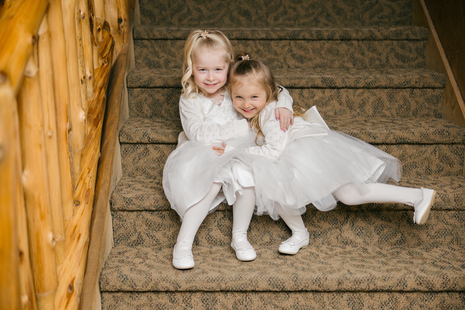 Two flower girls in white dresses sit hugging on a carpeted staircase beside a large log post inside Whitefish Lodge — Tim Larsen Photography, Brainerd Lakes MN