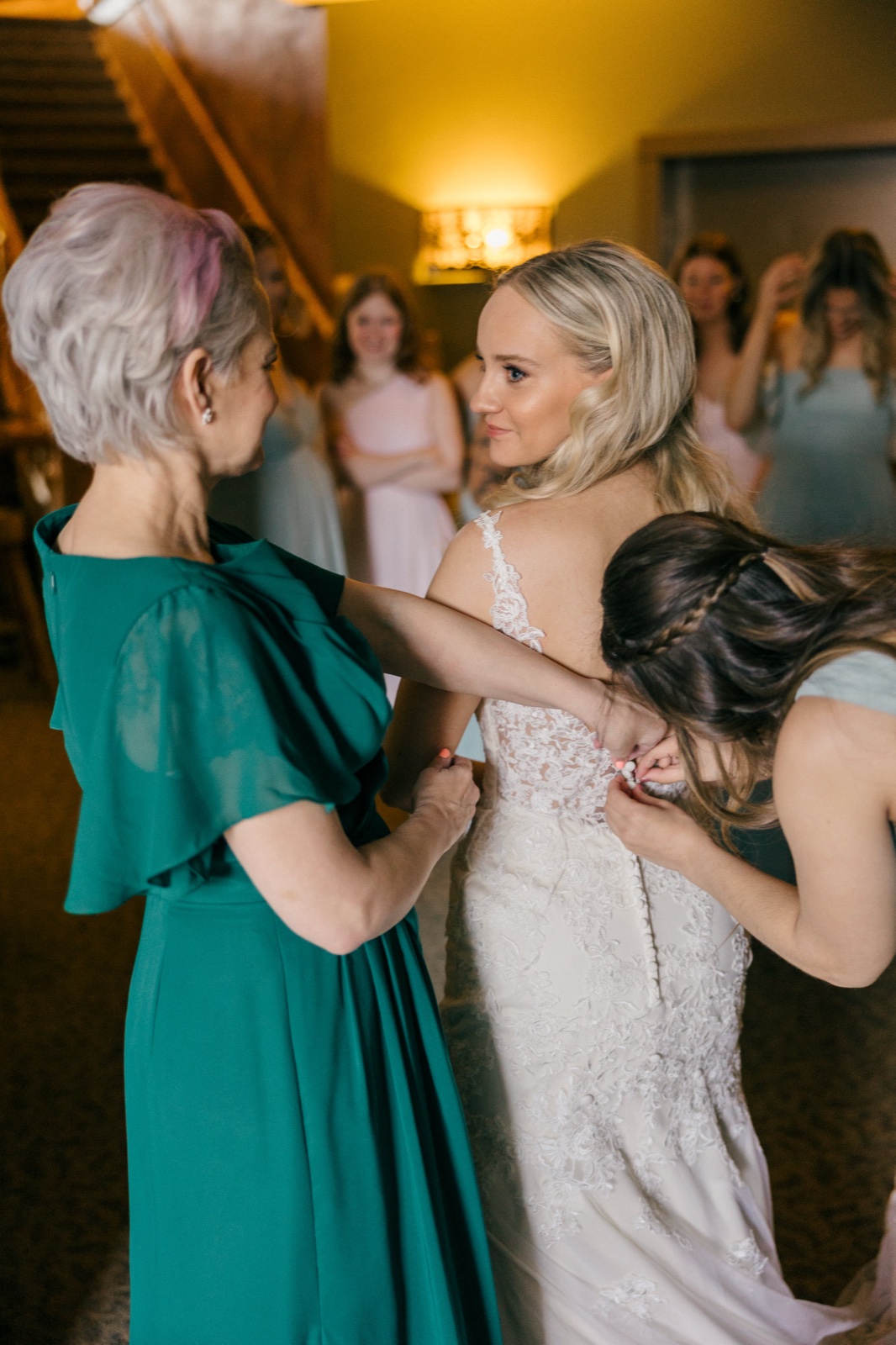 The bride stands between her mother in a teal dress and a bridesmaid buttoning the back of her lace gown, with other bridesmaids watching in the background — Tim Larsen Photography, Brainerd Lakes MN
