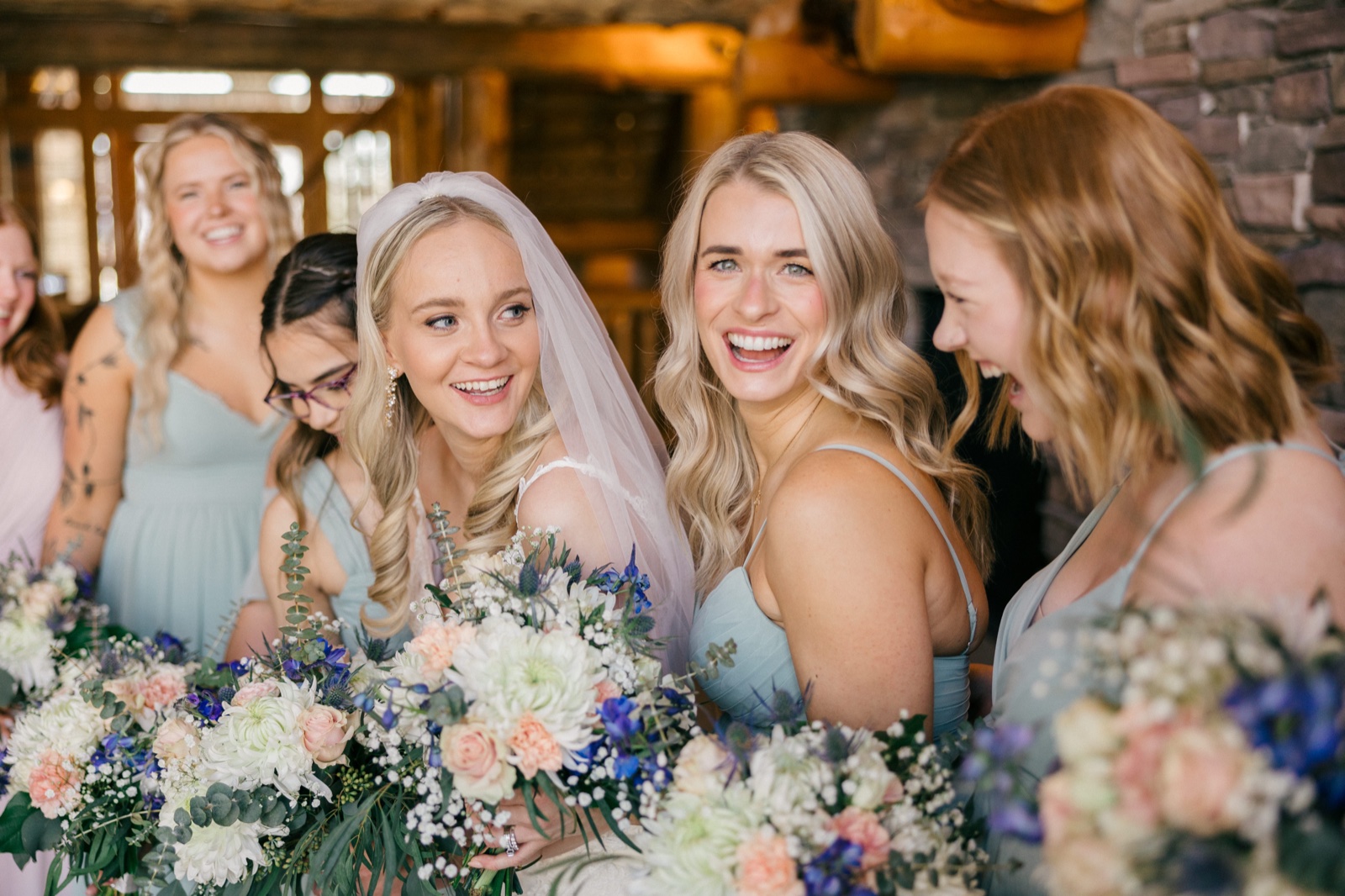 The bride in a veil smiles softly while her bridesmaids in dusty-blue dresses laugh together, holding pastel bouquets inside the wood-and-stone lodge — Tim Larsen Photography, Brainerd Lakes MN