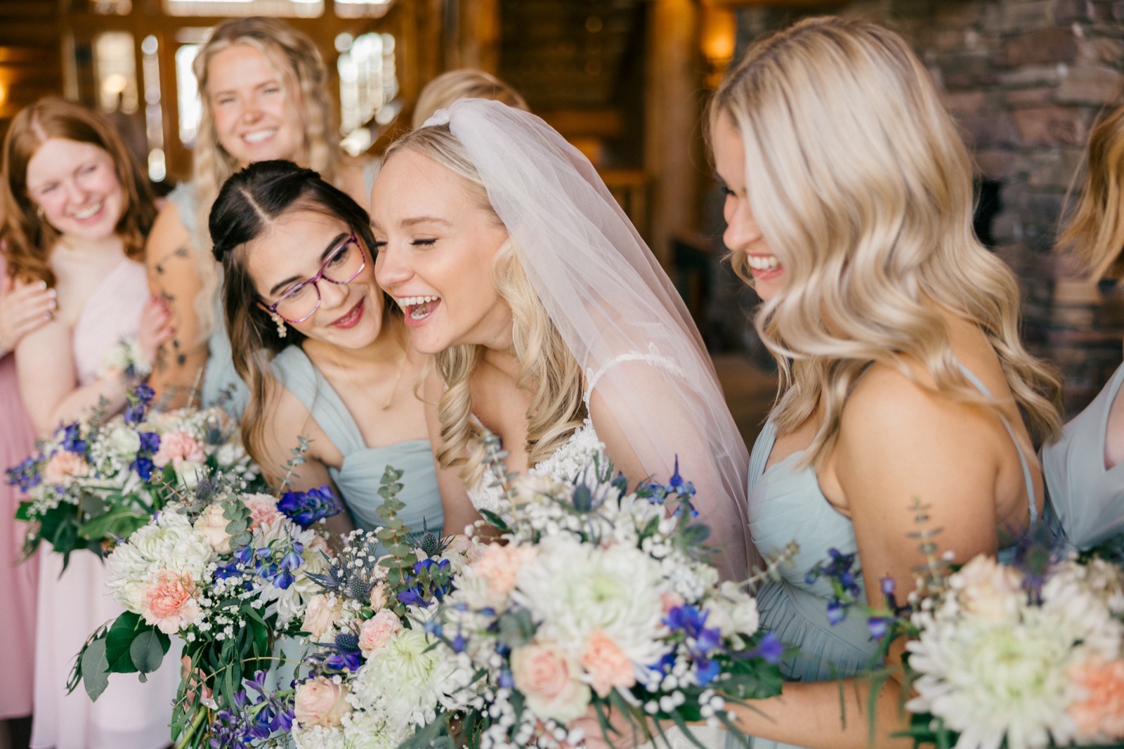 The bride laughs with her veil flowing back as a bridesmaid leans in close, both surrounded by other bridesmaids holding pastel bouquets — Tim Larsen Photography, Brainerd Lakes MN