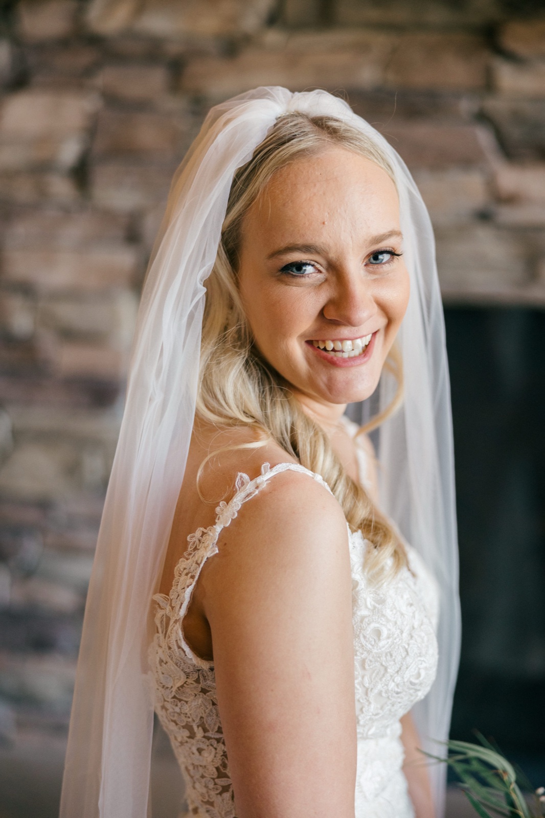 The bride smiles broadly over her shoulder, her veil draping down her back as she stands before a stone wall in soft window light — Tim Larsen Photography, Brainerd Lakes MN